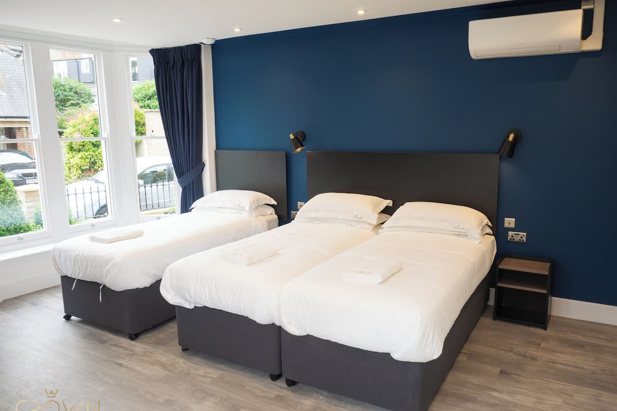 A hotel room with two beds, a large window with curtains, an air conditioning unit on the wall, and a small bedside table.