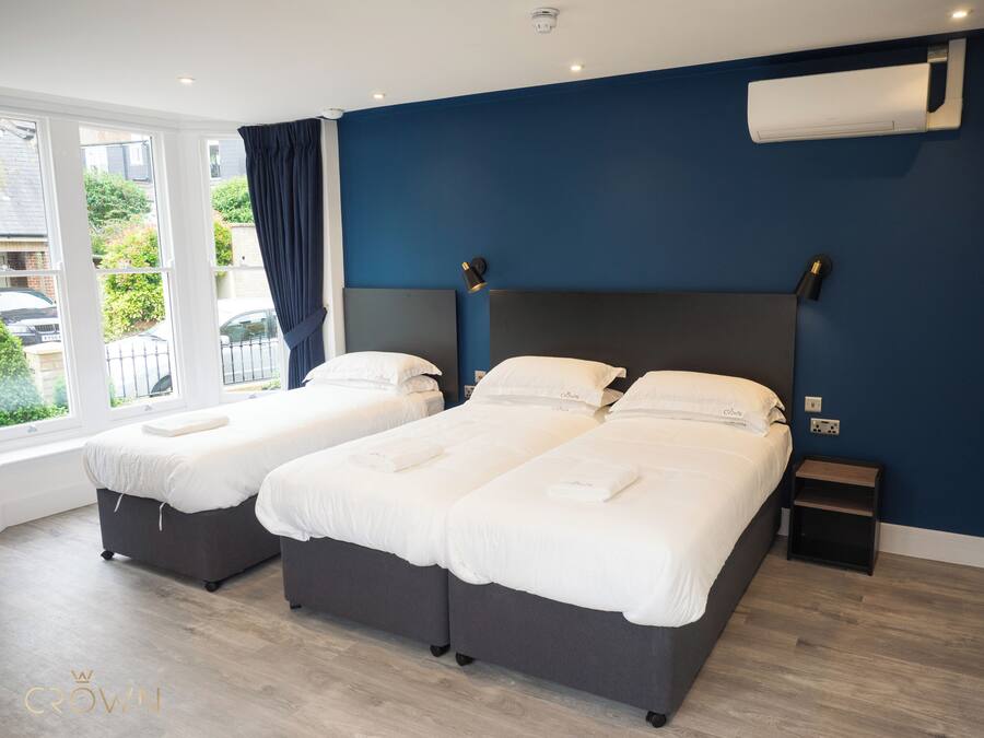 A hotel room with two beds, a large window with curtains, an air conditioning unit on the wall, and a small bedside table.