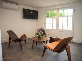 A hotel room with a round wooden table, two chairs, a flower vase, a flat-screen TV mounted on the wall, and a large window with a view of greenery.