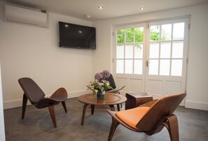 A hotel room with a round wooden table, two chairs, a flower vase, a flat-screen TV mounted on the wall, and a large window with a view of greenery. - Crown Hotel (Oxford)