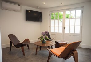 A hotel room with a round wooden table, two chairs, a flower vase, a flat-screen TV mounted on the wall, and a large window with a view of greenery.