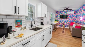 Cocina moderna con gabinetes blancos, grifo negro y protector contra salpicaduras de azulejos. Hay un área de comedor con un mural de colores llamativos, un televisor de pantalla plana y un sofá.