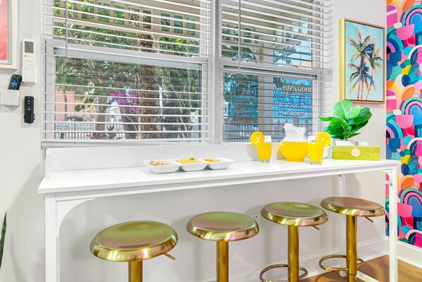 A kitchen with a white countertop, four gold stools, a window with blinds, and a colorful wall.