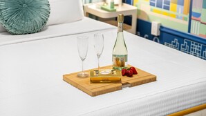 Una cama con una bandeja de madera que sostiene una botella de vino Verdi, dos copas y una caja de bombones Ferrero Rocher.