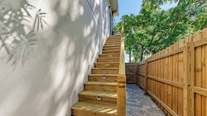 Una escalera de madera que sube hacia una pared blanca, con una cerca de madera y árboles al fondo.