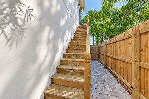 Eine Holztreppe führt zu einer weißen Außenwand, mit einem Holzzaun und einem gepflasterten Weg.