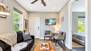 Una sala moderna con sofá, mesa de centro y dos sillones. Hay un televisor en la pared y una puerta que da a otra habitación.