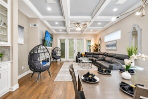 A modern living room with a sectional sofa, two black wireframe chairs, a dining table with a white plate and napkin, and a ceiling fan.