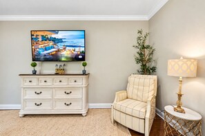 Un salon avec un téléviseur posé sur un meuble blanc, un fauteuil beige, une petite table avec une lampe et une plante.
