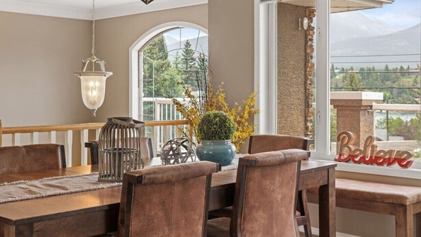 A dining area with a wooden table and chairs, a vase with yellow flowers, a hanging light fixture, and a view of a mountain through a large window. - Mountain Manor | Lake Views & Access | Fenced Yard (Invermere)