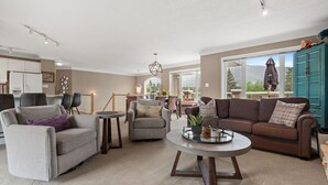 A spacious living room with a grey sofa, a brown sofa, and a round coffee table. There is a dining area with a small table and chairs. The room has a view of the outdoors through large windows. - Mountain Manor | Lake Views & Access | Fenced Yard (Invermere)