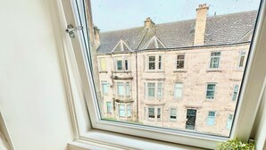 Une pièce avec une fenêtre donnant sur une rangée d’immeubles. On y trouve un panier en osier sur le sol et une plante en pot sur le rebord de la fenêtre.