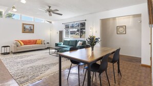 A spacious living room with a large window, a ceiling fan, a dining table with chairs, and a sofa with cushions. - Las Hermanas - Close to Sul ROss, Kokernot Park, Downtown (Alpine)