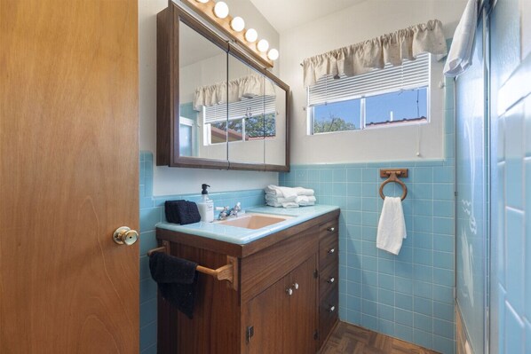 A bathroom with a wooden vanity, a sink, a mirror with lights, a towel rack, and a window with blinds.