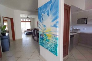 A spacious living area with a large painting of a seascape, a dining area with chairs, and a kitchen with modern appliances. - New Green Bay I Condos Golf View (Nuevo Nayarit)