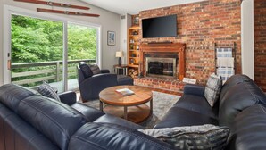 A modern living room with a brick fireplace, leather sofas, a flat-screen TV, and a balcony with a view of greenery.