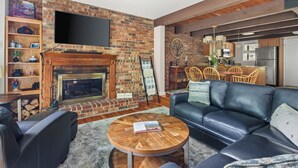 A living room with a brick fireplace, a flat-screen TV mounted above it, a leather sofa, a round coffee table, and a dining area with wooden chairs and a kitchen in the background.
