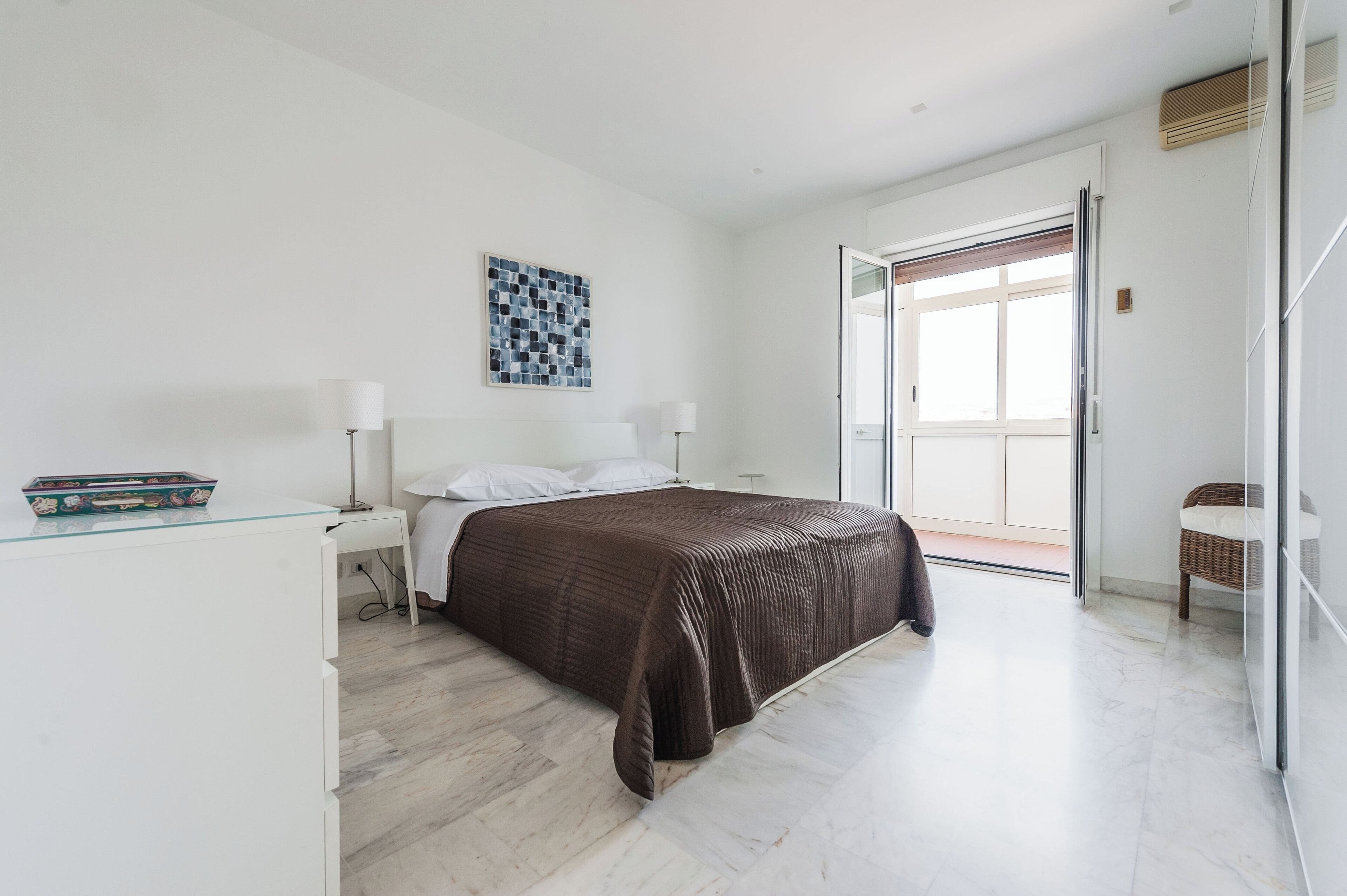 A modern bedroom with a bed, bedside tables, a lamp, a small table with a decorative item, and a view of the outside through a glass door.
