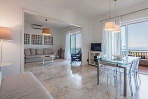 Un soggiorno moderno con divano, tavolo da pranzo in vetro e sedie trasparenti. Sul balcone si gode di una vista sul mare.