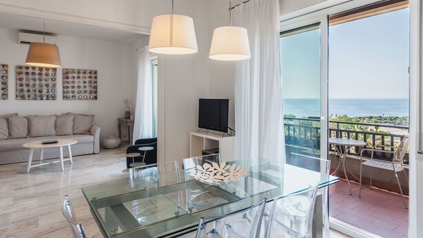 A modern living room with a glass dining table, a sofa, and a TV on a stand. There is a balcony with a view of the sea. - Europa Landscape by Wonderful Italy (Catania)