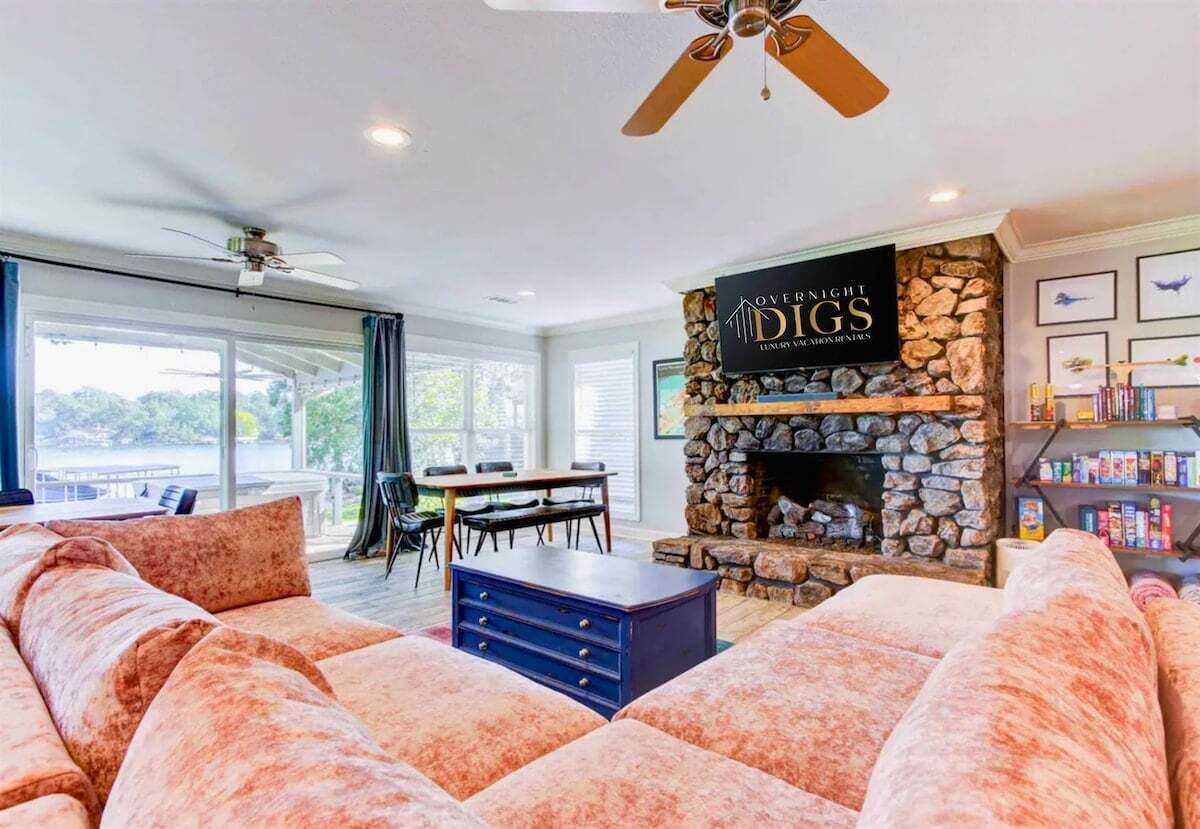 A spacious living room with a stone fireplace, a ceiling fan, and a large window overlooking a deck.