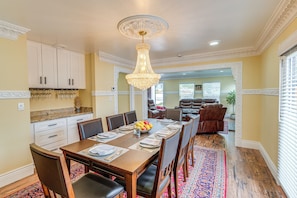A dining area with a wooden table set for six, surrounded by chairs. There is a chandelier above the table, and the room has cream-colored walls and a patterned rug on the floor.