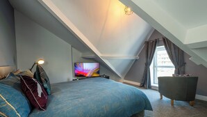 Une chambre avec un plafond en pente, un lit recouvert d’une couverture bleue, une télévision, une chaise et une fenêtre avec des rideaux.