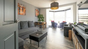 Un salon moderne avec un canapé gris, une table basse, un coin repas avec des chaises et un espace cuisine doté d’un comptoir et d’appareils électroménagers.