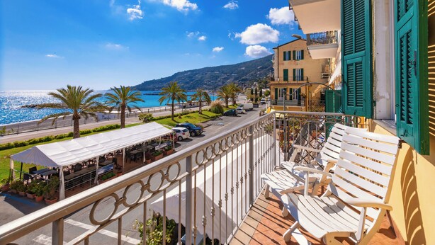A balcony with white chairs, a view of the sea, and a building with green shutters. - Appartamento fronte mare a Ospedaletti by Wonderful Italy (Ospedaletti)