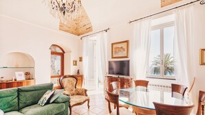 A living room with a green sofa, a glass dining table, and wooden chairs. There's a TV on a wooden cabinet, and a chandelier hangs from the ceiling. - Appartamento fronte mare a Ospedaletti by Wonderful Italy (Ospedaletti)