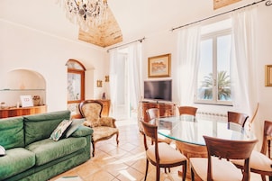 Una sala de estar con un sofá verde, una mesa de comedor de cristal y sillas de madera. Hay un televisor sobre un mueble de madera, y un candelabro cuelga del techo.
