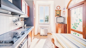 A compact kitchen with a stove, sink, and a small refrigerator. There is a window with a view of palm trees and a door with a wooden frame. - Appartamento fronte mare a Ospedaletti by Wonderful Italy (Ospedaletti)