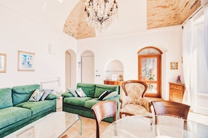 Un salón con un sofá verde, una silla de madera, una mesa de centro de vidrio y una araña.