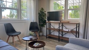 Un salon moderne comprenant un canapé, deux fauteuils, une table basse, un téléviseur posé sur un meuble en bois et de grandes fenêtres avec des rideaux.
