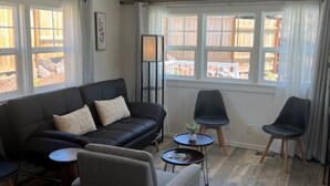 Un salon moderne avec un canapé en cuir noir, un fauteuil gris et deux chaises noires. On y trouve deux petites tables rondes, un ventilateur de plafond et de grandes fenêtres avec des rideaux.