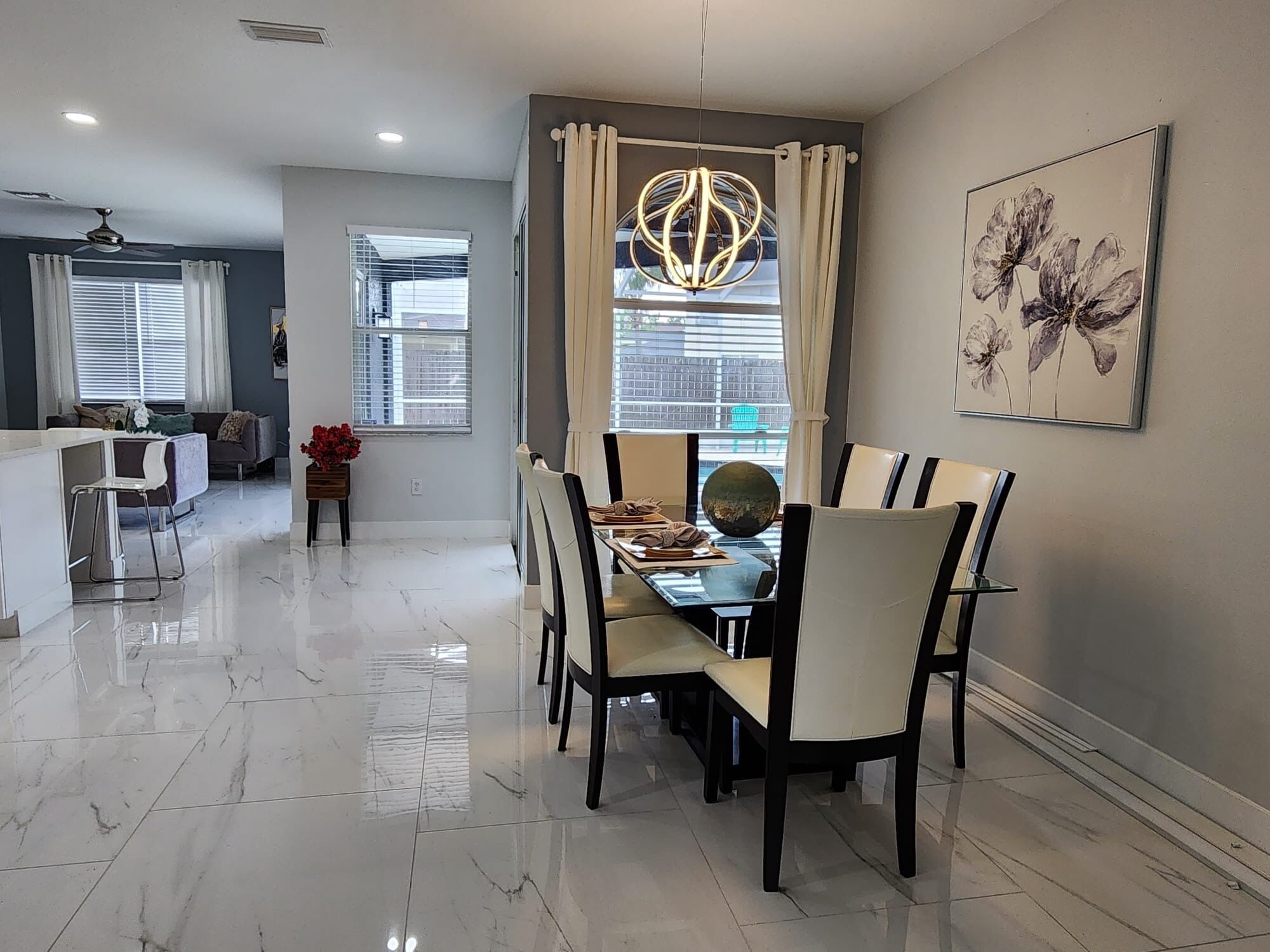 A modern dining area with a marble floor, a dining table with chairs, a chandelier, and a large window with curtains.