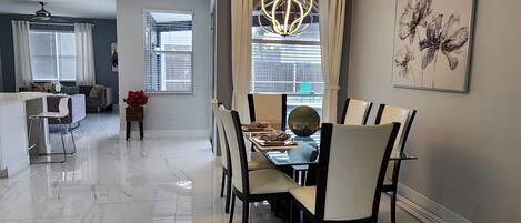 A modern dining area with a marble floor, a dining table with chairs, a chandelier, and a large window with curtains.