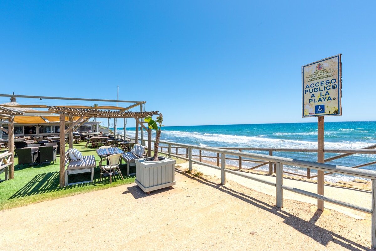 A beachside restaurant with outdoor seating, a sign indicating public beach access, and a view of the ocean.
