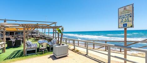 A beachside restaurant with outdoor seating, a sign indicating public beach access, and a view of the ocean.
