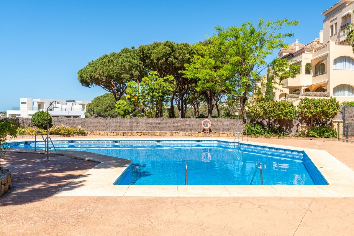 A swimming pool with a clear blue water, surrounded by a paved area, trees, and a building with balconies.