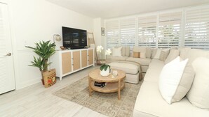 A living room with a beige sofa, a round coffee table, a television on a cabinet, and a large window with blinds.
