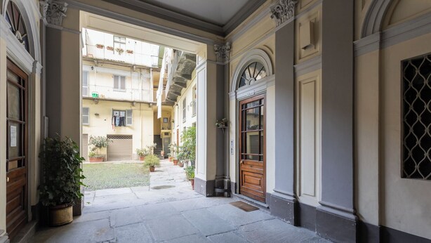 A stone-paved courtyard with a wooden door labeled "SCHOLA," surrounded by buildings with balconies and potted plants. - Casa Juliette by Wonderful Italy (Turin)