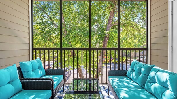 A covered porch with two teal sofas and a small table, overlooking a street with trees and parked cars. - Newly Renovated Condo Minutes from The Beach, OWA Water Park and Tanger Outlets (Foley)