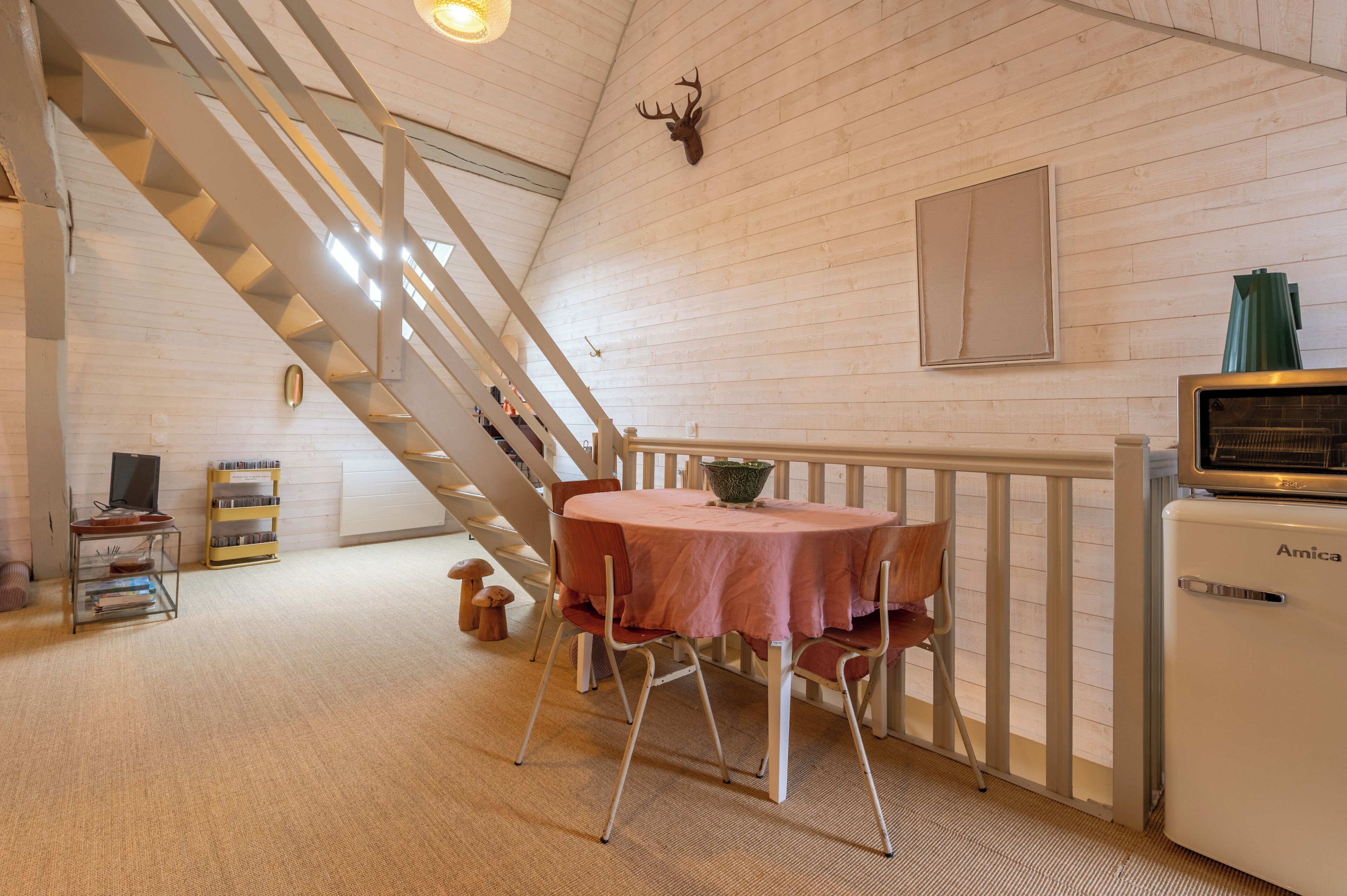 Een kamer met een trap, een ronde tafel met stoelen, een kleine koelkast en een opgeplakte hertenkop aan de muur.