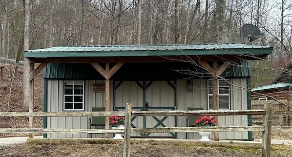 A rustic cabin with a green roof and wooden beams, surrounded by a wooden fence and a small garden with red flowers. - Turkey Ridge Lodges- Cabin 10 (Rockbridge)