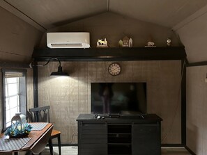 Una habitación con techo de madera, aire acondicionado instalado, un escritorio con lámpara, un reloj y un televisor en la pared.