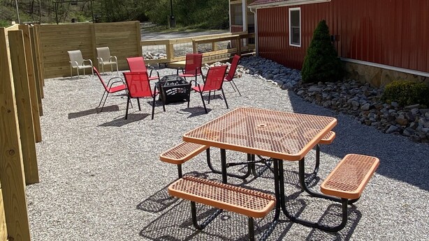 An outdoor seating area with a picnic table and chairs, a wooden deck, and a red building in the background. - Turkey Ridge Lodges - The Nest (ROCKBRIDGE)