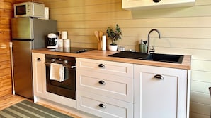 A compact kitchen with wooden panel walls, a stainless steel refrigerator, a built-in oven, a microwave, a sink, and a small potted plant. - Chalet de la Prairie (Le Hom)