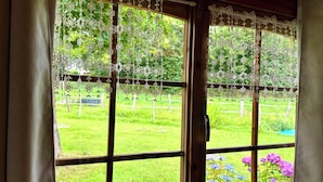 A window with lace curtains and a view of a grassy yard with flowers. - Chalet de la Prairie (Le Hom)