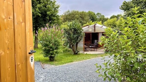 A rustic wooden door leading to a gravel path, a small wooden structure with a covered patio, and lush greenery. - Chalet de la Prairie (Le Hom)